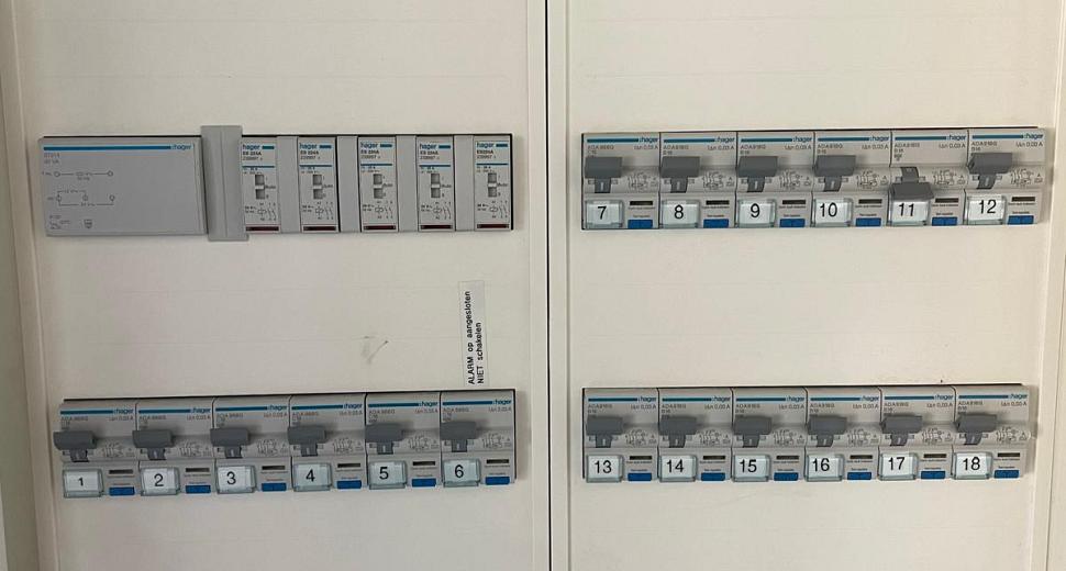 Close-up van een elektriciteitskast met Waasdorp-logo’s, hoofdschakelaar en genummerde stroomgroepen voor gebouwbeheer, netjes geordend en gelabeld.