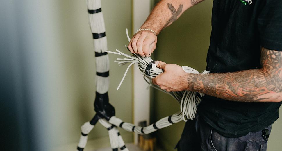 Man in werkkleding bundelt witte kabels met zwarte tape tegen een groene muur. De kabels zijn deels aan de muur bevestigd.