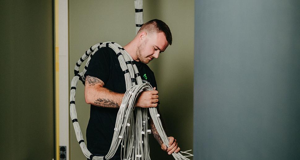 Technicus draagt een grote bundel kabels over zijn schouder in een binnenruimte met groene muren, klaar voor installatie.