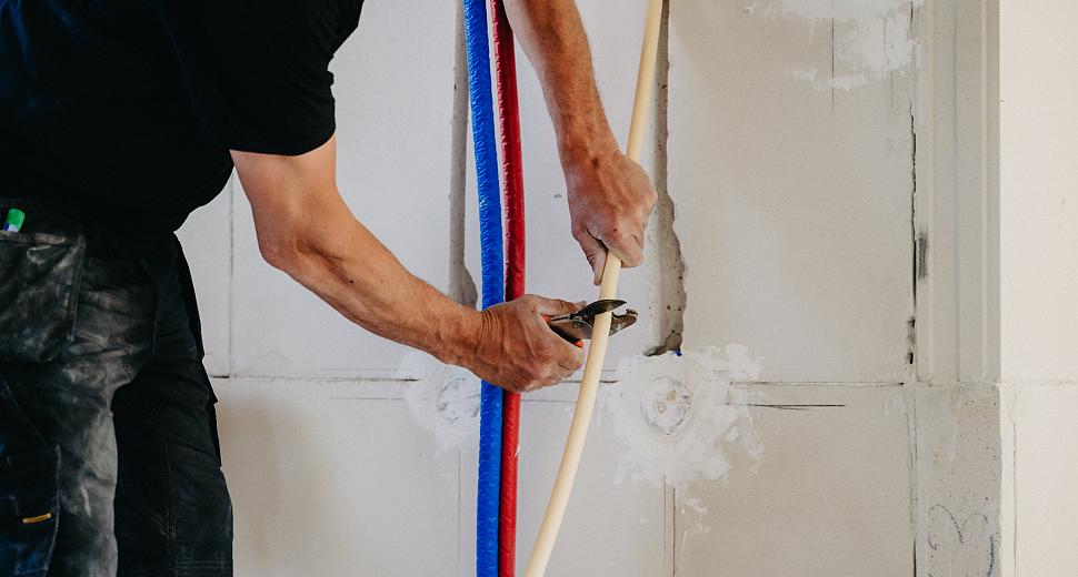 Elektricien snijdt een elektriciteitsbuis op maat tegen een muur met sleuven voor kabelinstallatie, met zicht op blauwe en rode mantelbuizen.