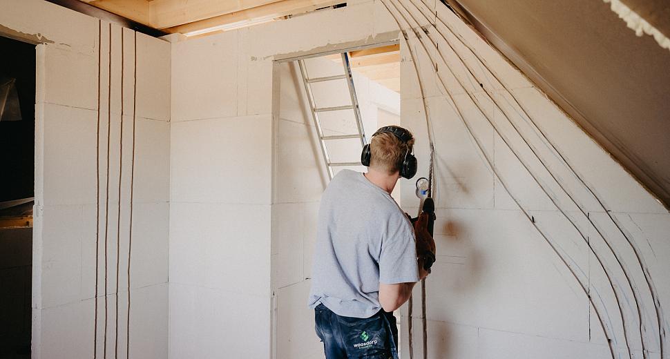 Elektricien maakt sleuven in een muur van een nieuwbouwwoning met een muurfrees, om elektriciteitsleidingen te plaatsen. Veiligheidsuitrusting zoals gehoorbescherming zichtbaar.