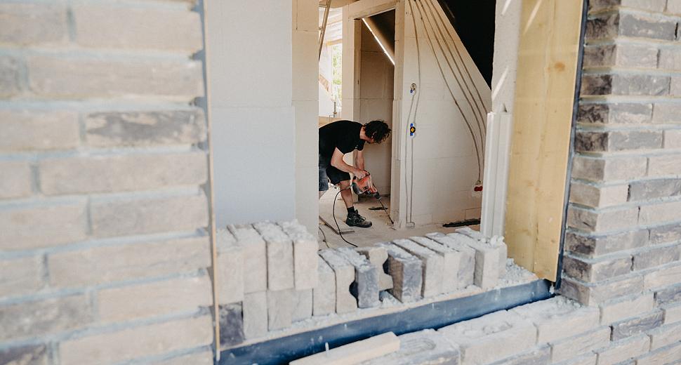 Bouwvakker aan het werk in een nieuwbouwwoning, zichtbaar door een onafgewerkt raam. Hij maakt sleuven in een binnenmuur voor elektrische bedrading.