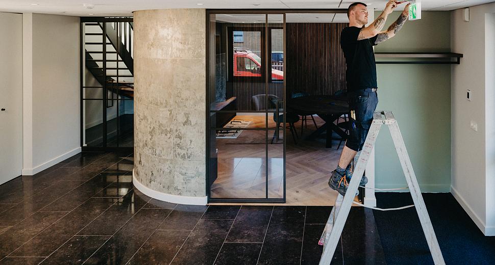  Monteur op een ladder die een noodverlichting monteert aan het plafond in een modern kantoorinterieur met glazen deuren, natuurstenen vloer en betonnen muur.
