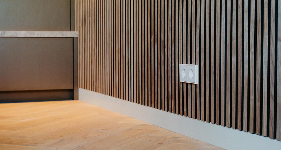 Houten wand met verticale lamellen in een moderne interieursetting, met een dubbel stopcontact geïntegreerd. De vloer is van licht hout in visgraatmotief.