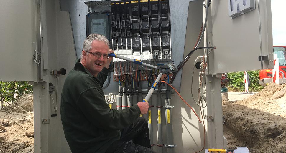 Monteur lachend bezig met het aansluiten van een groot elektrisch verdeelstation buiten, omringd door gereedschap en opengewerkte bekabeling.