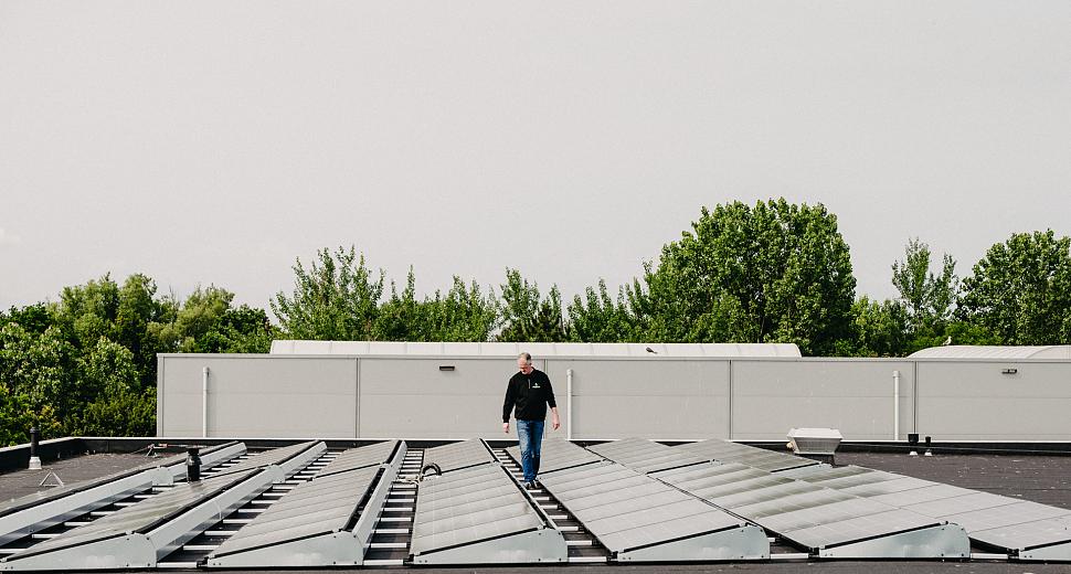 Man loopt over een plat dak met geïnstalleerde zonnepanelen en montagesystemen, omgeven door bomen en een witte muur op de achtergrond.