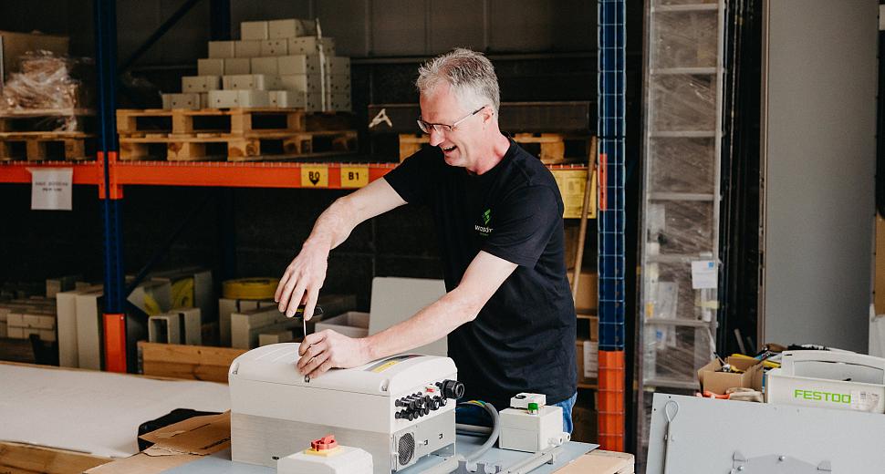 Een technicus werkt aan een elektrische installatiekast op een houten werkbank in een magazijnomgeving, omringd door rekken met apparatuur en materialen.