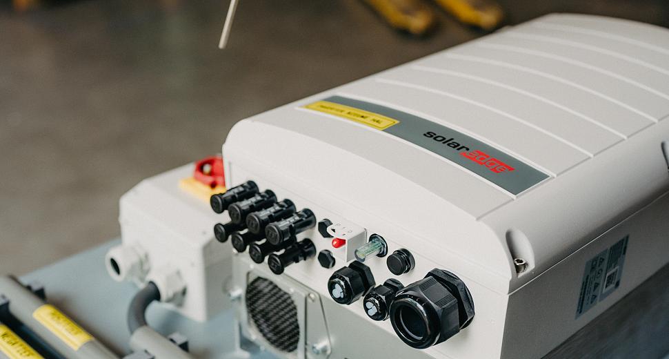 Close-up van een SolarEdge omvormer met diverse aansluitingen en bedrading, geplaatst op een werkblad in een magazijnomgeving.