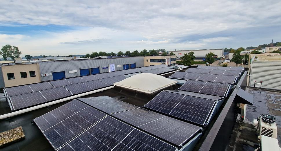 Zonnepanelen geïnstalleerd op een plat dak met een lichtkoepel in het midden, omringd door industriële panden en een bewolkte lucht op de achtergrond.