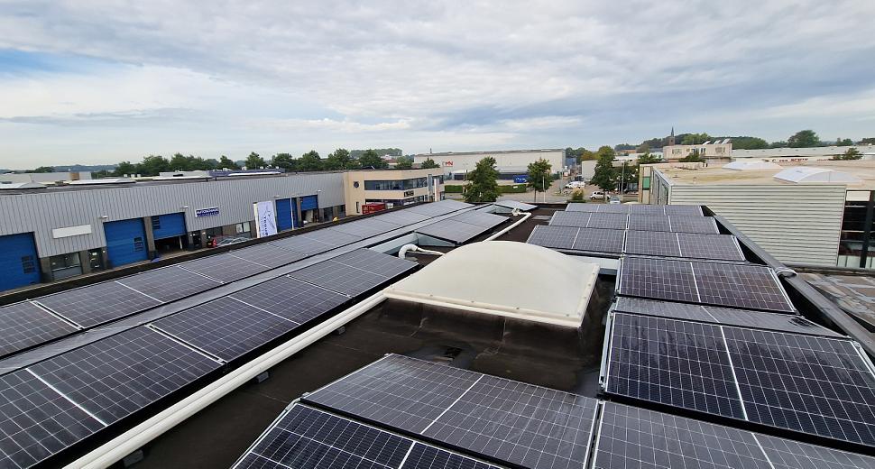 Zonnepanelen geïnstalleerd op een plat dak met een lichtkoepel in het midden, omringd door industriële panden en een bewolkte lucht op de achtergrond.
