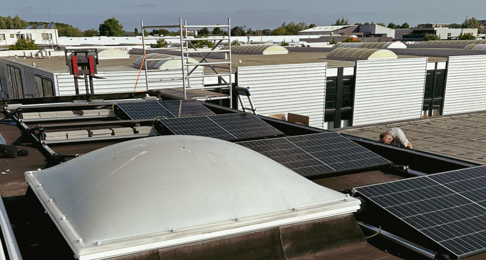 Plat dak met geïnstalleerde zonnepanelen rondom een lichtkoepel, met op de achtergrond een steiger en industriële gebouwen. Een persoon werkt aan de panelen.
