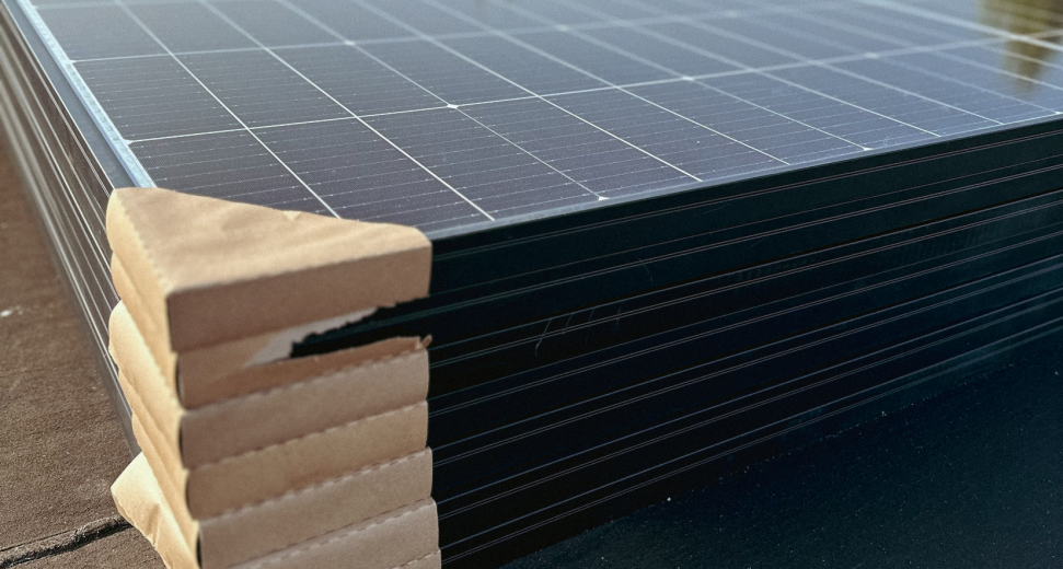 Stapel nieuwe zonnepanelen, strak verpakt in karton, geplaatst op een plat dak, klaar voor installatie. Reflectie van zonlicht op de panelen.