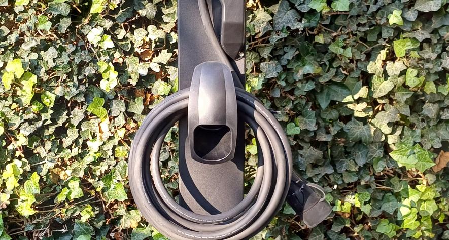  Donkergrijze elektrische laadpaal met opgerolde laadkabel, geplaatst tegen een achtergrond van groene klimop en stenen bestrating.