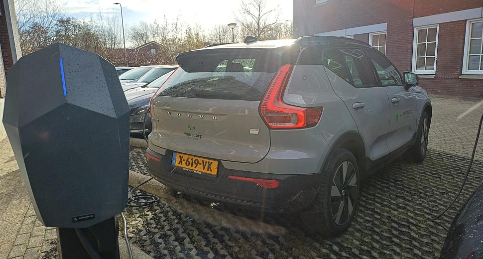Elektrische laadpaal verbonden met een grijze Volvo XC40 met Waasdorp-logo, geparkeerd op een oprit bij een bakstenen gebouw onder een heldere hemel.