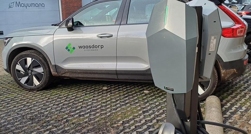 Elektrische laadpaal met opgerolde kabel, geplaatst op een parkeerplaats. Op de achtergrond een auto met het logo van Waasdorp E-Techniek en een bakstenen gebouw.