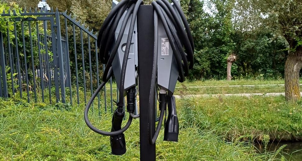 Zwarte elektrische laadpaal met opgerolde laadkabels, geplaatst in een groene omgeving met bomen, struiken en een hekwerk op de achtergrond.