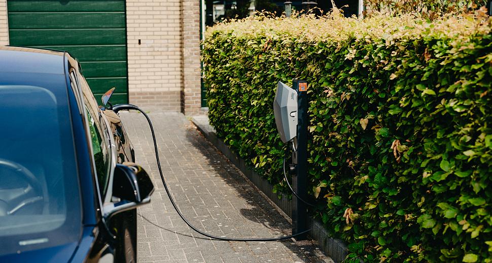 Elektrische auto aangesloten op een laadpaal naast een groene heg, gelegen op een oprit voor een huis met een groene garagedeur op de achtergrond.