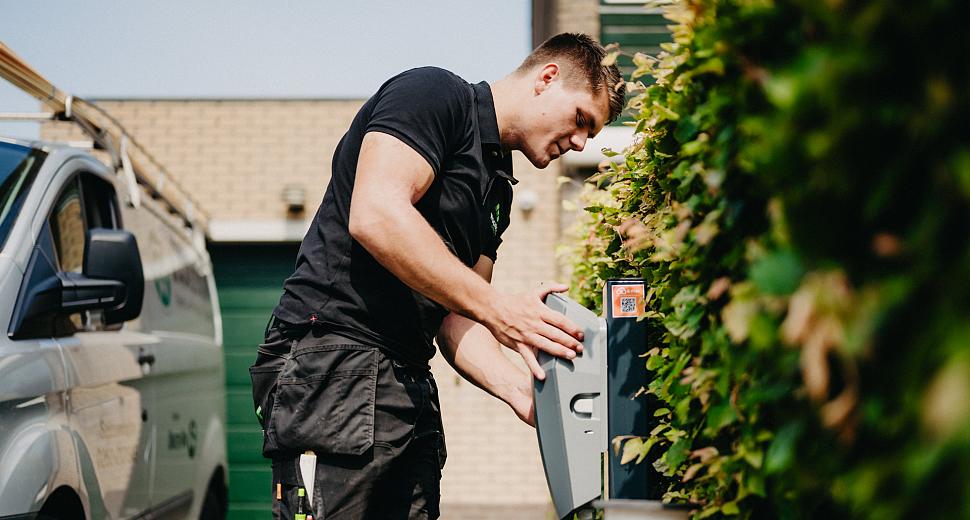  Monteur in werktenue installeert een laadpaal naast een groene heg, met een busje en een huis met groene garagedeur op de achtergrond.