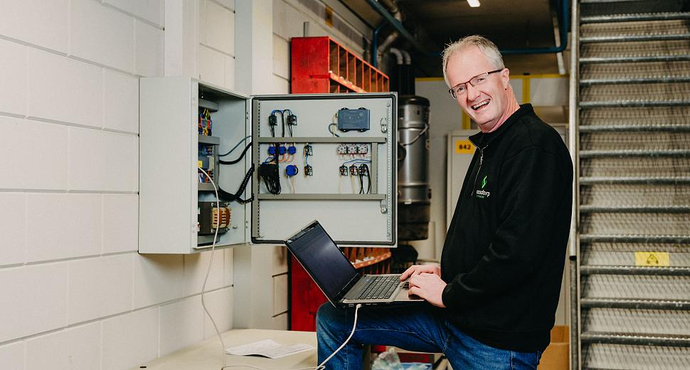 Lachende technicus met een laptop werkt aan een geopende besturingskast in een industriële ruimte, met zicht op trappen en technische apparatuur.