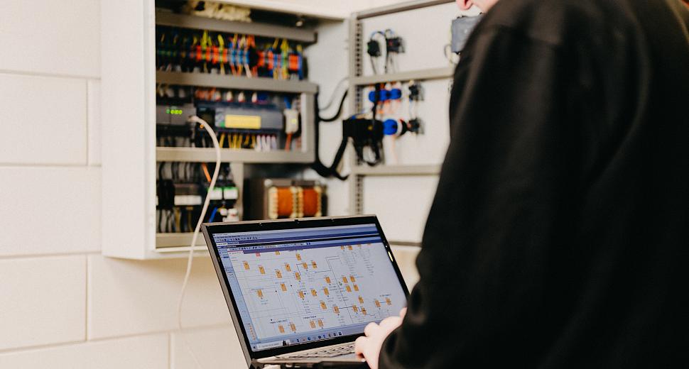 Technicus werkt met een laptop aan een geopende schakelkast, met een zichtbaar digitaal schema op het laptopscherm en bedrading in de kast.