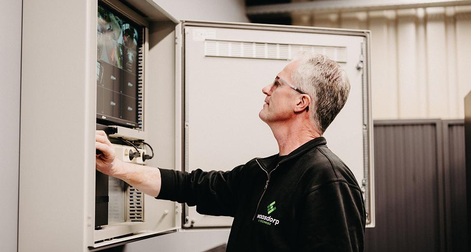 Medewerker van Waasdorp E-Techniek bedient een monitor in een open schakelkast, gekleed in een zwart bedrijfstrui, binnen een industriële omgevin