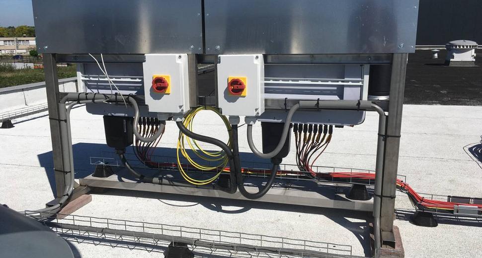  Twee metalen omvormers gemonteerd op een stalen frame op een plat dak, met aangesloten kabels en elektriciteitsvoorzieningen, tegen een blauwe lucht.