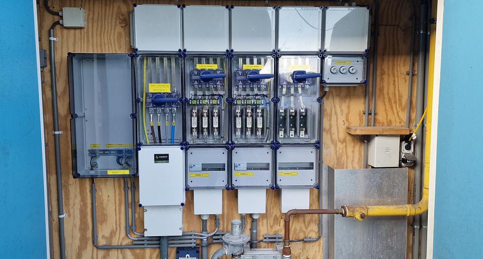 Elektrische verdeelkast met diverse meters en schakelaars, gemonteerd op een houten achtergrond in een blauwe behuizing, met zichtbare gasleiding rechts.