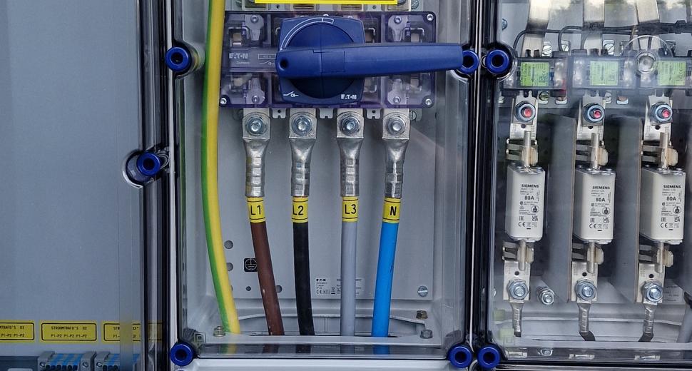  Close-up van een elektrische schakelkast met een doorzichtig paneel, zichtbare bedrading en een gele hoofdschakelaar, gemonteerd op een houten wand.