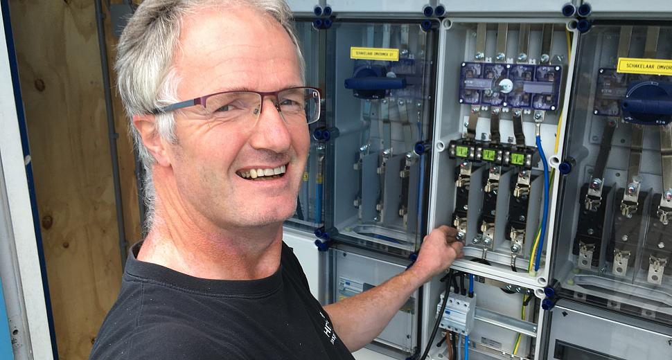 Monteur met veiligheidsbril bij een geopende elektrische schakelkast met zichtbare componenten en bedrading, werkend aan onderhoud of installatie.