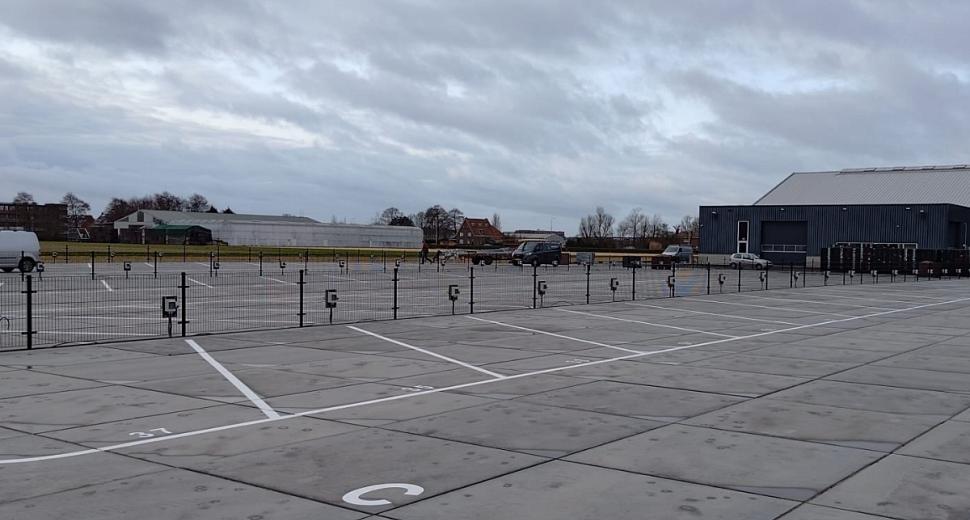 Parkeerterrein met genummerde vakken, omheind door metalen hekken. Op de achtergrond staan enkele gebouwen en voertuigen onder een bewolkte lucht.