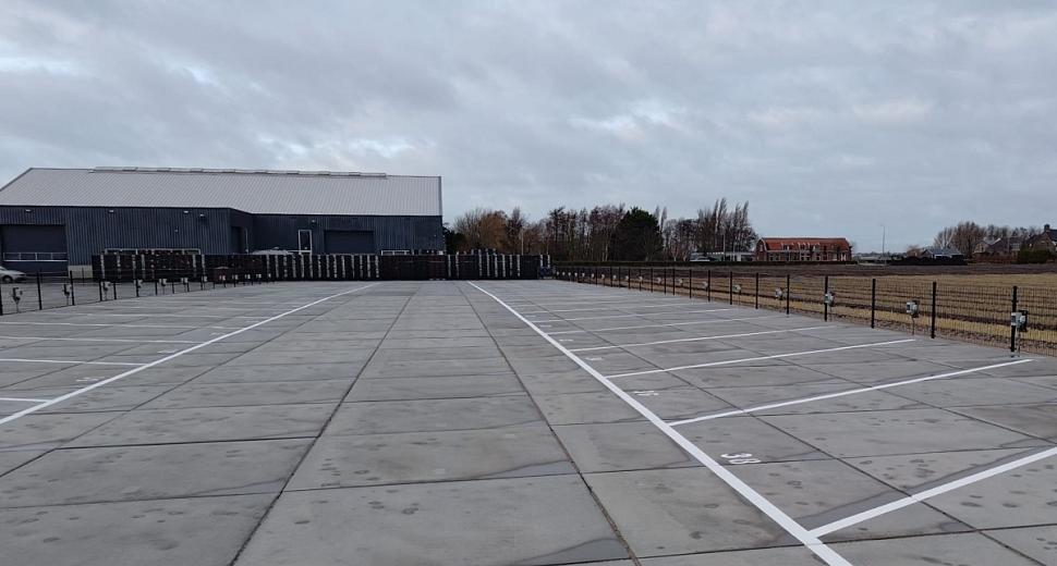  Betegeld parkeerterrein met genummerde vakken en metalen omheining. Op de achtergrond staat een groot grijs gebouw, omlijst door bomen en een bewolkte lucht.