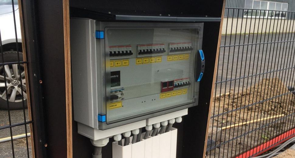 Elektriciteitskast in een houten frame met een doorzichtige deur, zichtbaar vanaf de zijkant. Labels en schakelaars zijn duidelijk te zien. Op de achtergrond een hek, auto en gebouw.