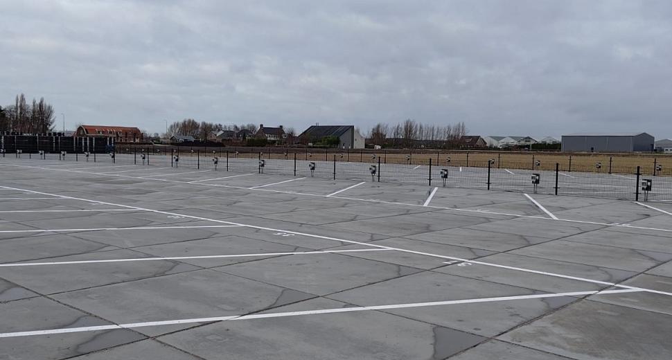 Grote open parkeerplaats met witte belijning en oplaadpunten voor elektrische voertuigen langs een hek. Op de achtergrond huizen en lage bebouwing.