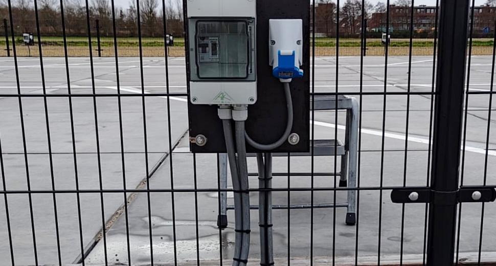 Oplaadpunt voor elektrische voertuigen bevestigd aan een metalen hek, met een stroomaansluiting en kabels zichtbaar. Achtergrond toont een parkeerplaats.