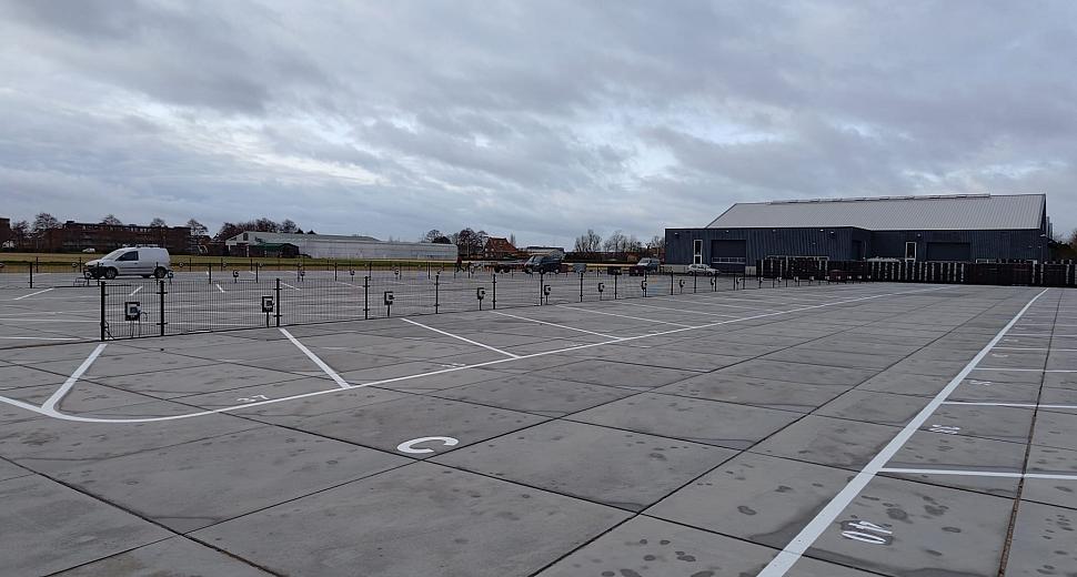 Parkeerterrein met genummerde vakken, omheind door metalen hekken. Op de achtergrond staan enkele gebouwen en voertuigen onder een bewolkte lucht.