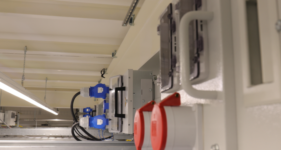 Detail van een technische installatie met rode en blauwe industriële aansluitingen, bevestigd aan een witte structuur, met metalen kabelgoten en verlichting op de achtergrond.