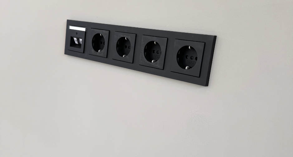 Zwarte wandmodule met vier stopcontacten en één netwerkaansluiting, strak geïntegreerd in een lichtgrijze muur voor een moderne uitstraling.