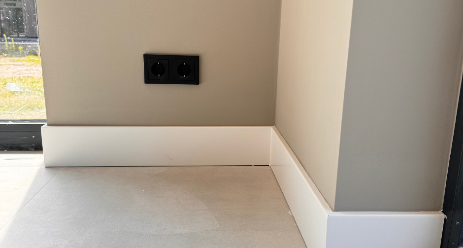 Hoek van een kamer met een lichtgrijze muur, witte plinten en een zwarte dubbele wandcontactdoos, naast een raam met zicht op een tuin.