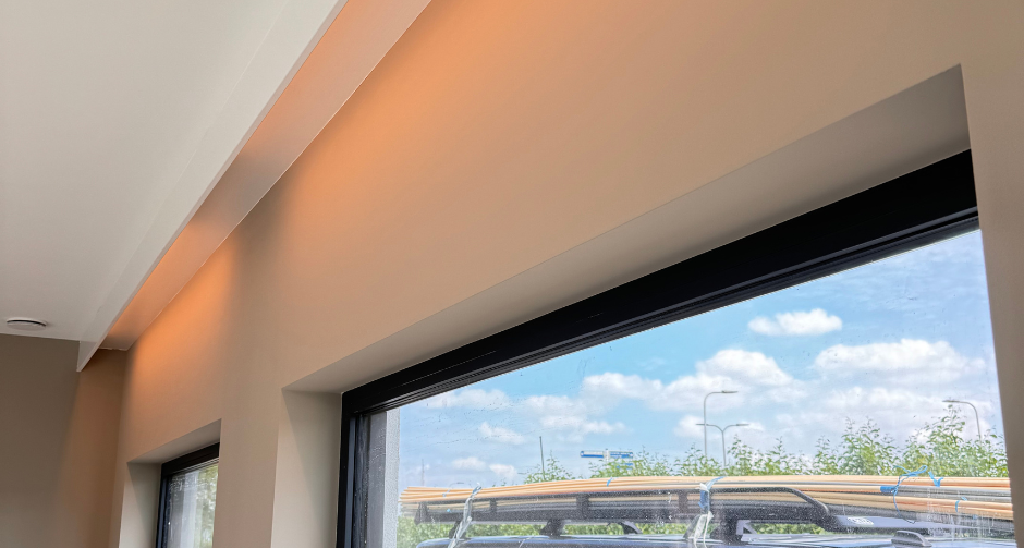 Ramen met zwarte kozijnen in een moderne ruimte, met zachte indirecte verlichting bovenaan de muur. Buiten is een bedrijfswagen met "Waasdorp E-techniek" zichtbaar.