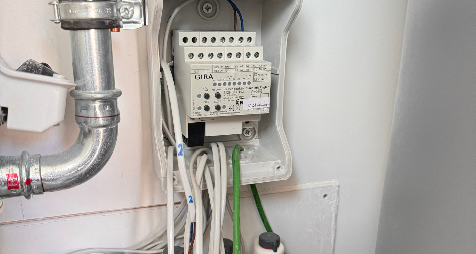 Open schakelkast met een GIRA KNX-module, aangesloten op meerdere kabels en leidingen. Onderin zijn verwarmingsverdeleronderdelen met actuators zichtbaar.