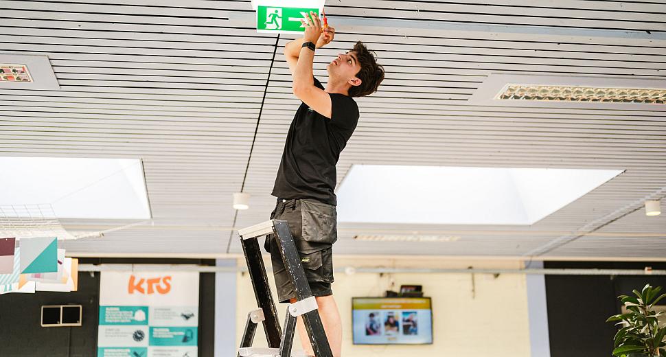 Technicus op een ladder in een binnenruimte, bezig met de installatie of het onderhoud van een groen nooduitgangbord aan het plafond.