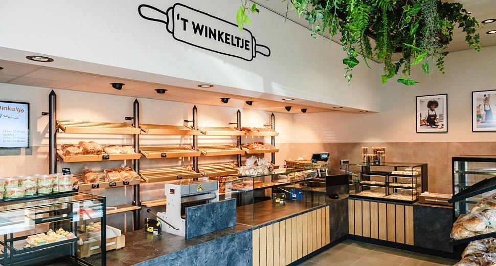 Interieur van een moderne bakkerij genaamd 't Winkeltje, met houten en stenen afwerking, gevuld met vers brood en gebak, onder een plafond met groene planten.