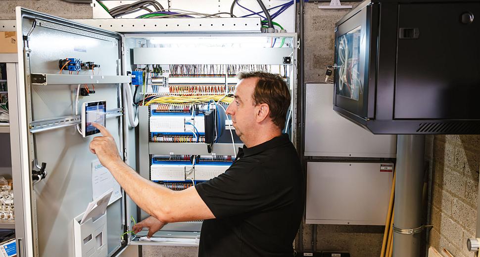 Technicus werkt aan een open schakelkast vol bedrading en apparatuur, in een industriële omgeving met zichtbaar leidingwerk en een monitor aan de muur.