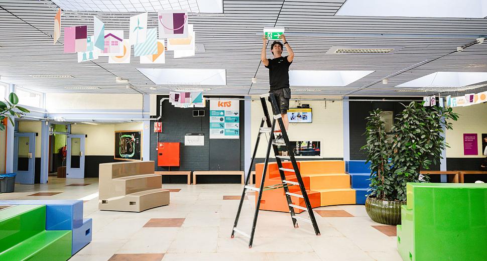 Technicus op een ladder in een open schoolruimte, bezig met het installeren of controleren van een nooduitgangbord aan het plafond. Omringd door kleurrijke decoraties en meubels.
