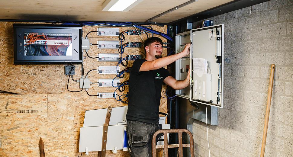 Technicus controleert een open schakelkast aan een houten wand met bedrading en technische apparatuur, terwijl een monitor bovenaan het systeem is aangesloten.