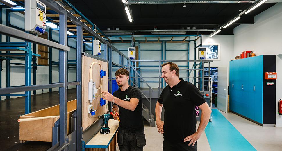 Een jonge monteur van Waasdorp E-Techniek werkt aan een elektrisch paneel, begeleid door een ervaren collega in een werkplaats met blauwe kasten en gereedschap.
