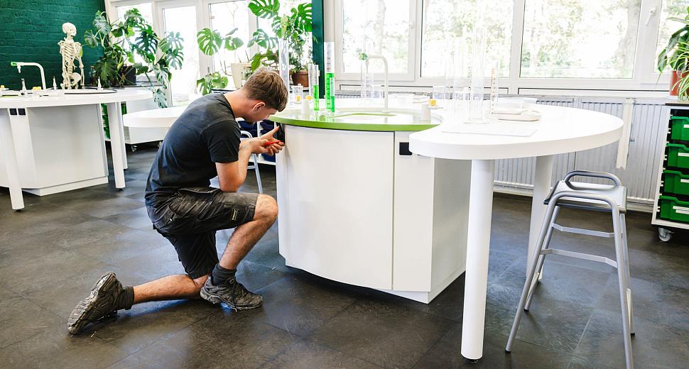 Een monteur die knielt om een kastje te repareren in een klaslokaal, omringd door werktafels, planten en grote ramen met uitzicht op een groene omgeving.