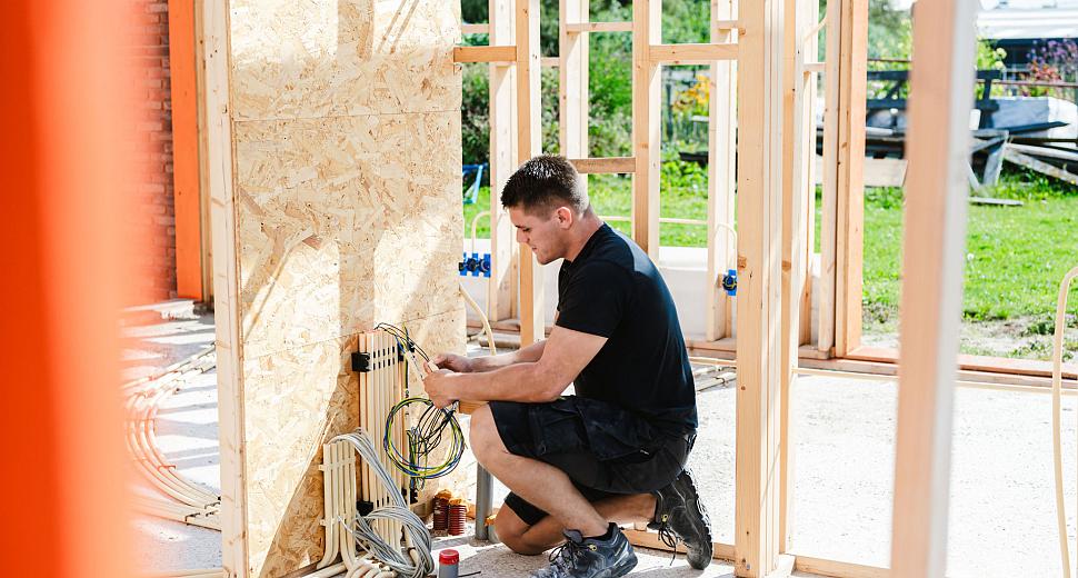 Elektricien werkt aan elektrische bedrading in een houten skeletbouw, met zicht op buizen en kabels tegen een OSB-muur en een groene achtergrond.