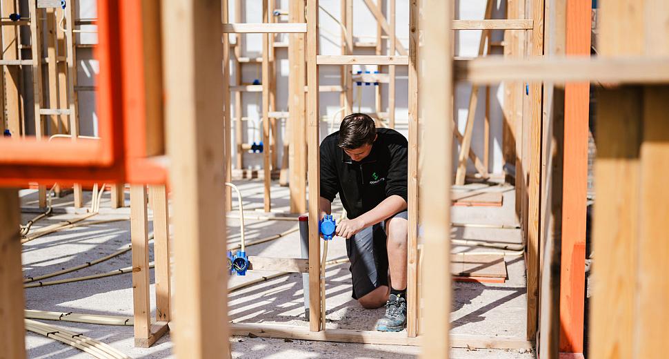 Monteur installeert blauwe inbouwdozen in een houten skeletbouwconstructie, omgeven door houten stijlen en zichtbare elektrische leidingen.