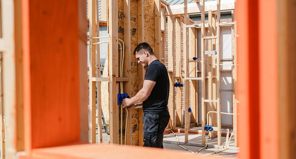 Monteur installeert blauwe inbouwdozen in een houten skeletbouwconstructie, gefotografeerd door een oranje kader, met zichtbare leidingen en een zonnige achtergrond.